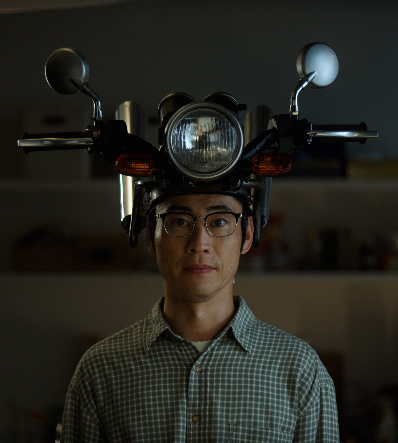 バイクを頭に載せて、自宅でこちらを見つめるチェックのシャツの男性